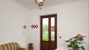 A room with a dining table set for four, a sofa, a decorative vase, and a view of a garden through a door. - Stunning Farmhouse in Passignano With Pool (Passignano sul Trasimeno)