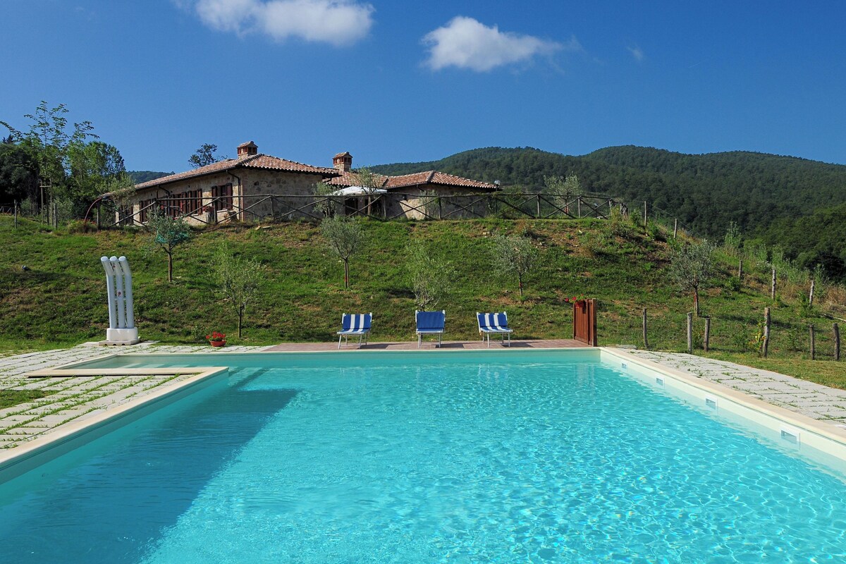 Una piscina con sillones de descanso, un edificio de piedra con techo rojo y un paisaje montañoso al fondo.