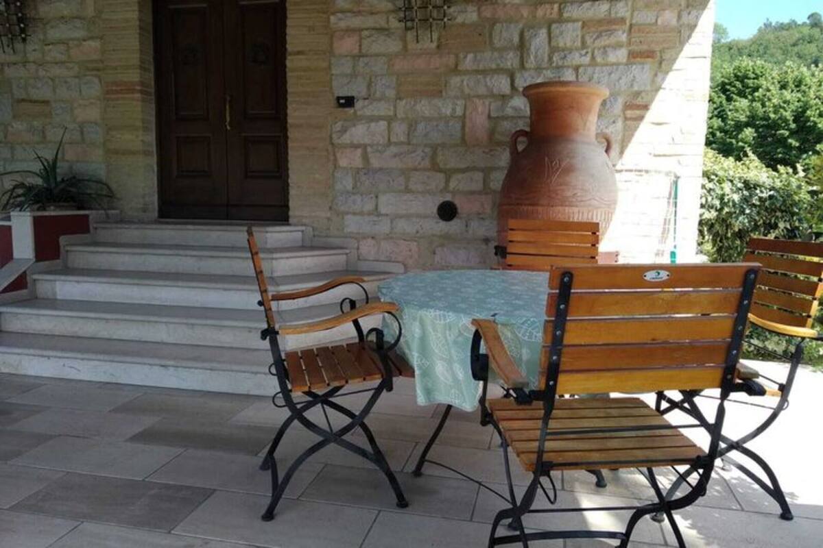A stone building with a wooden door, a large terracotta pot, and a table with chairs outside.