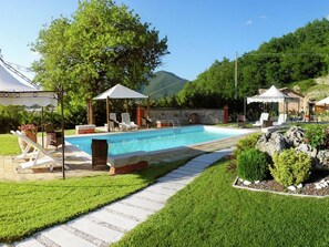 Una piscina con sillones de descanso y sombrillas, rodeada de césped cuidado y un sendero de piedra.
