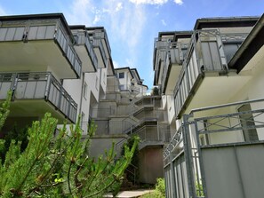 A multi-story residential building with balconies, staircases, and a small garden area with pine trees.