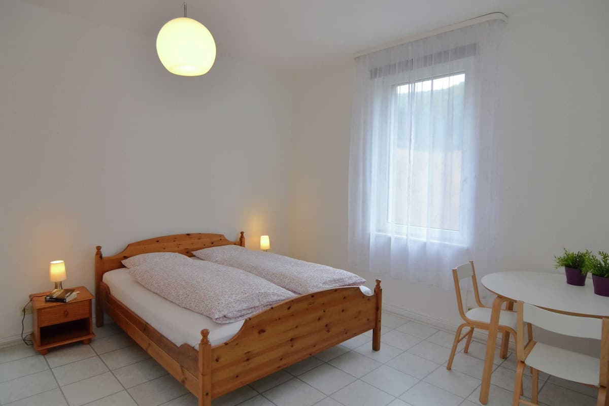 Een slaapkamer met een houten bed, nachtlampen, een ronde tafel met stoelen en een raam met dunne gordijnen.