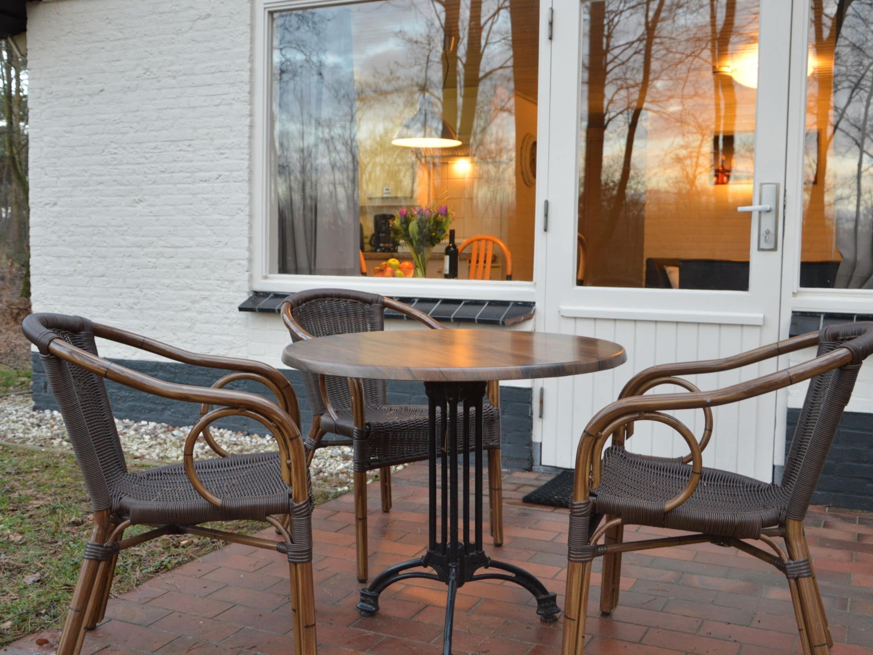 Een terras met rieten stoelen en een ronde tafel, een witte buitenmuur en een raam met uitzicht op bomen en een lamp van binnen.