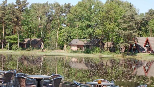 A lakeside outdoor seating area with wicker chairs and tables, surrounded by trees and buildings. - Completely Detached Bungalow in a Nature-filled Park by a Large fen (Stramproy)