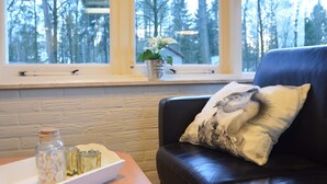 A room with a leather sofa, a cushion with a bird design, a window with a view of trees, and a table with a jar and glasses. - Completely Detached Bungalow in a Nature-filled Park by a Large fen (Stramproy)