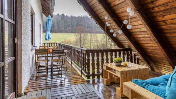 Een houten terras met een houten tafel en stoelen, een blauwe bank, en uitzicht op een bebost gebied.