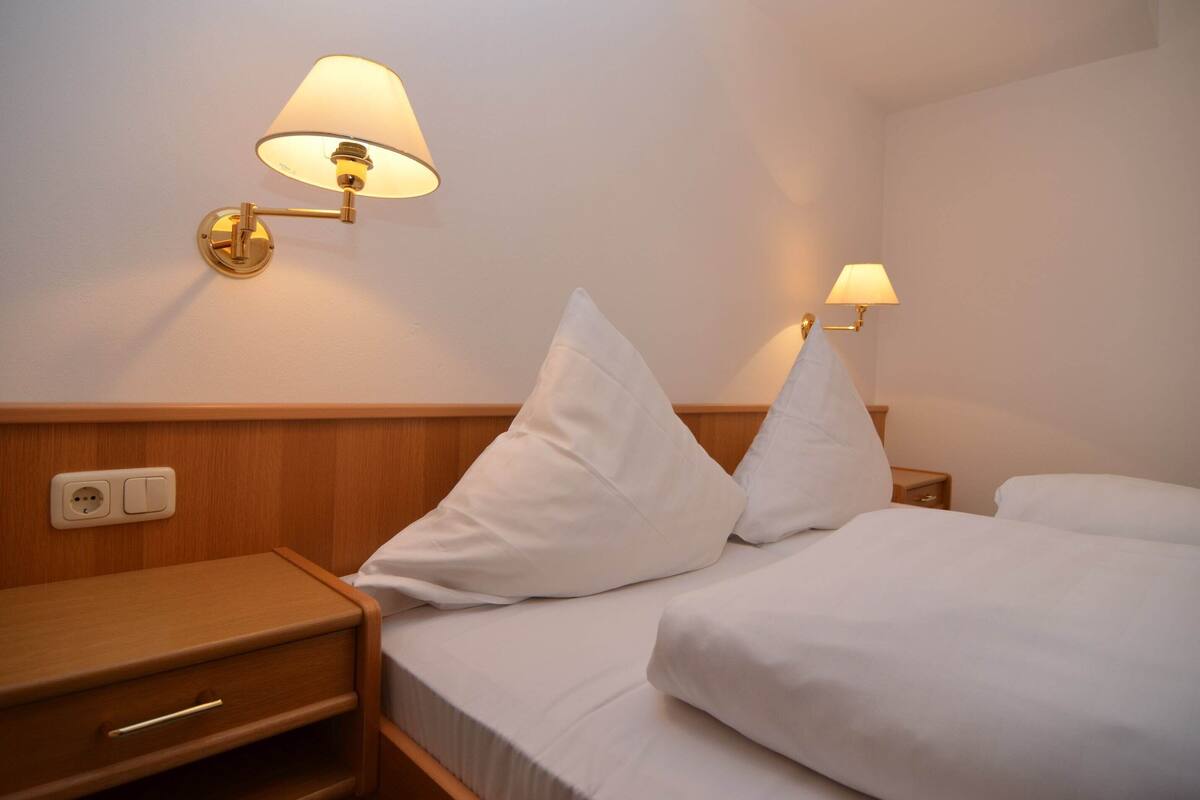 Une chambre d’hôtel avec deux lits, chacun doté de draps et d’oreillers blancs, une table de chevet en bois avec un tiroir et deux lampes fixées au mur.