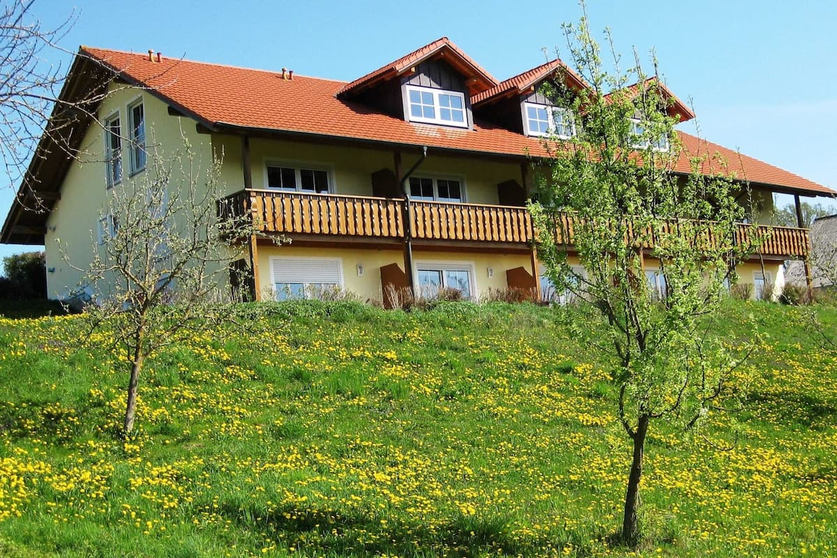 A two-story house with a red-tiled roof, multiple balconies, and a grassy lawn with yellow flowers.