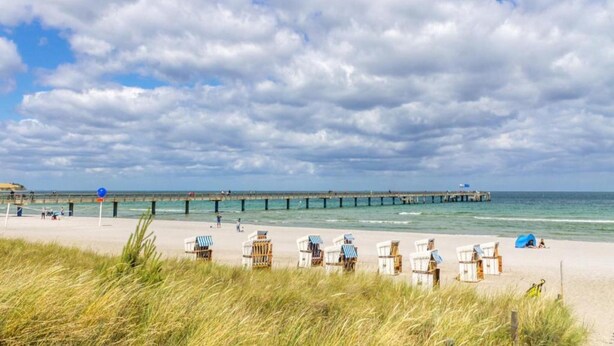 Ein Strand mit einem hölzernen Steg, der ins Meer führt, Strandliegen in Reih und Glied und einem wolkenlosen Himmel.