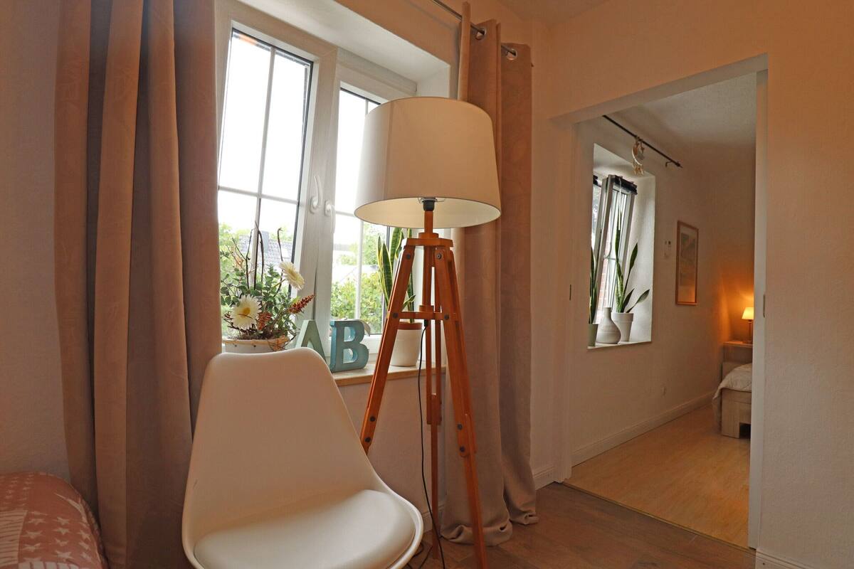 A room with a white chair, a floor lamp, a window with curtains, and a view of another room.