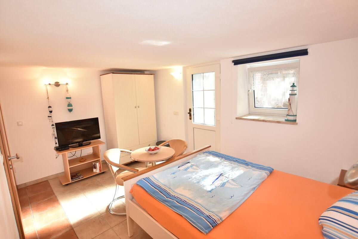 A small, well-lit room with a bed, a desk with a television, a chair, and a window with a curtain.