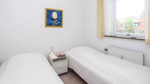 A bedroom with two single beds, a bedside table with a lamp, a framed picture on the wall, and a window with a view of buildings. - 4 Person Holiday Home in Allinge (Allinge)