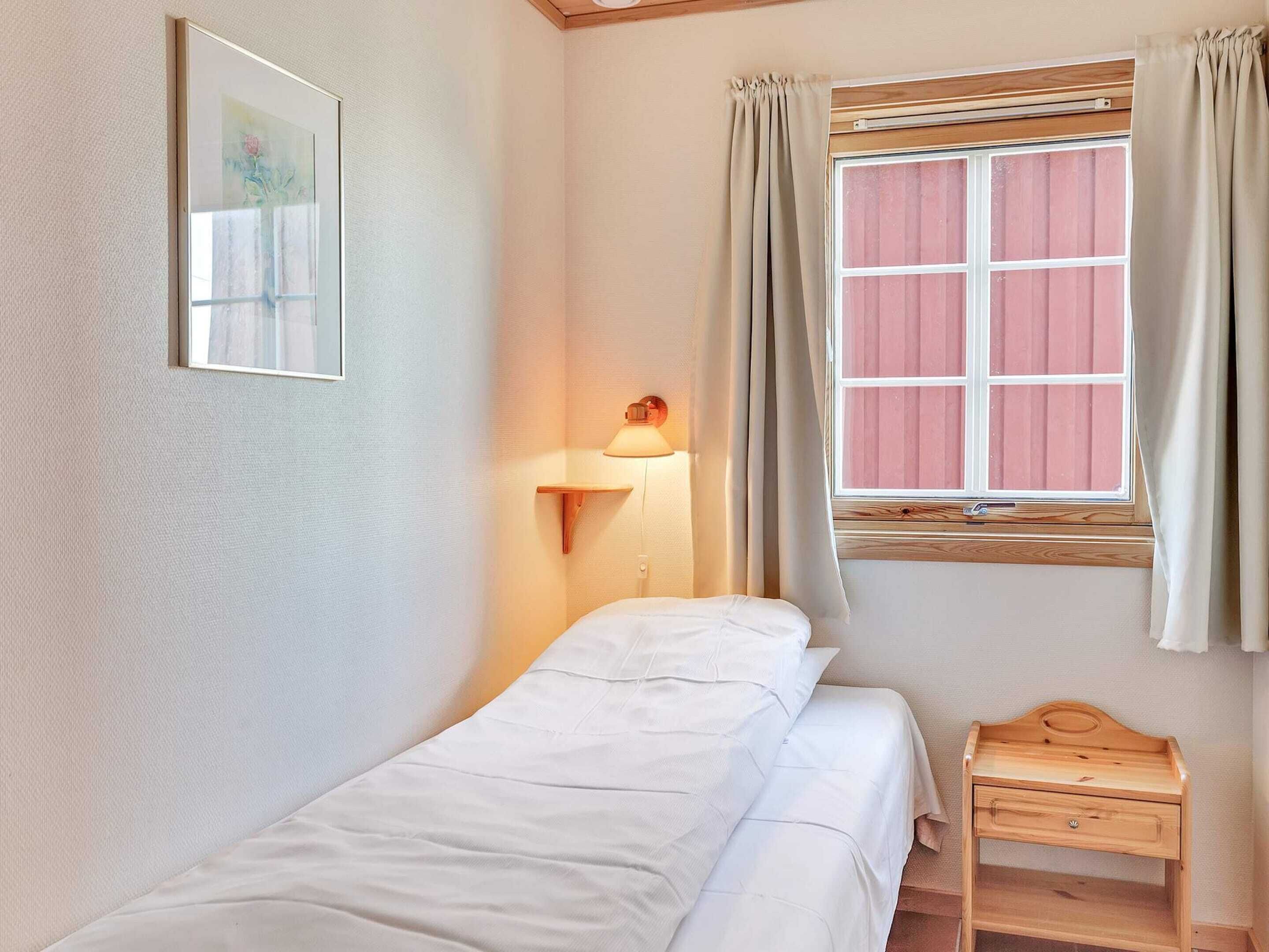 A small, neatly made bedroom with a single bed, a bedside table, a lamp, and a window with curtains.