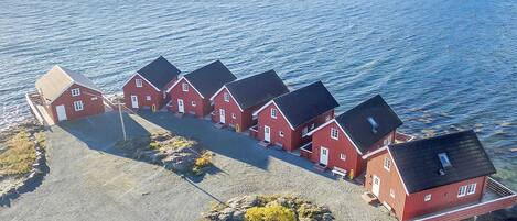 Una hilera de casas de madera rojas con techos negros, ubicadas en una costa rocosa con vista al mar y montañas distantes.