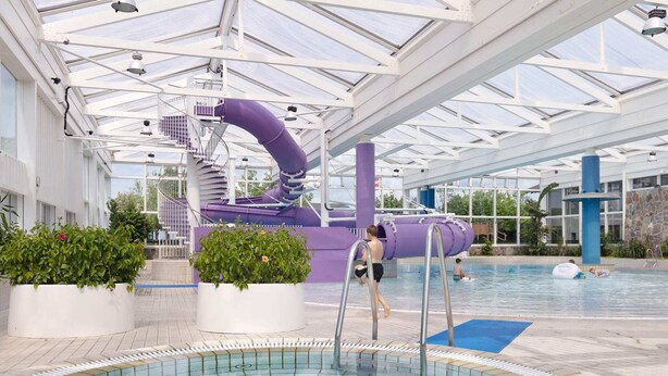 An indoor swimming pool with a slide, surrounded by potted plants and a covered area with a glass roof.