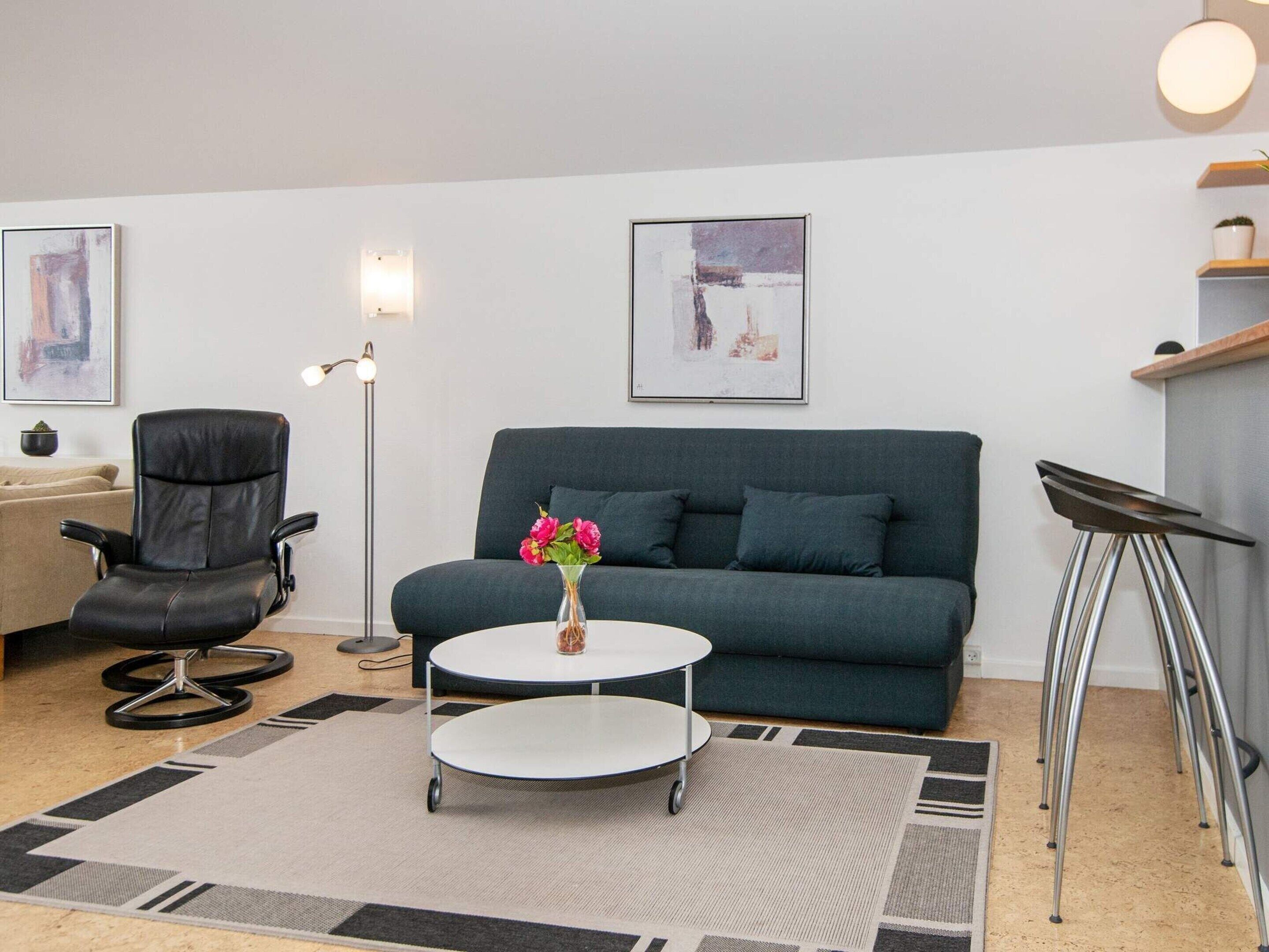 A modern living room with a grey sofa, a black office chair, a white coffee table with a vase of flowers, and a floor lamp.