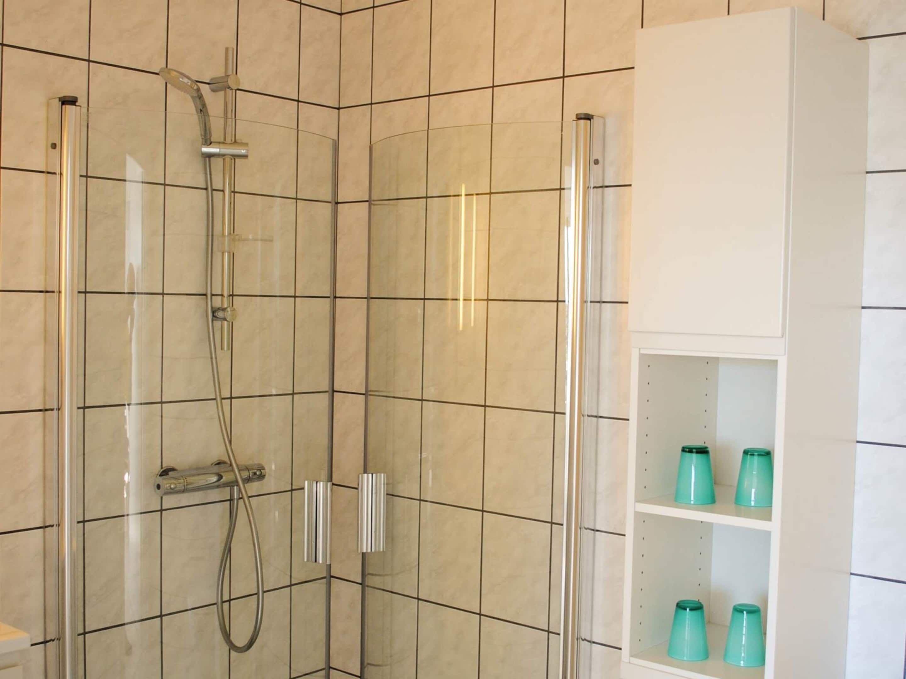 A modern bathroom with a glass-enclosed shower, beige tiled walls, and a white cabinet with teal bottles.