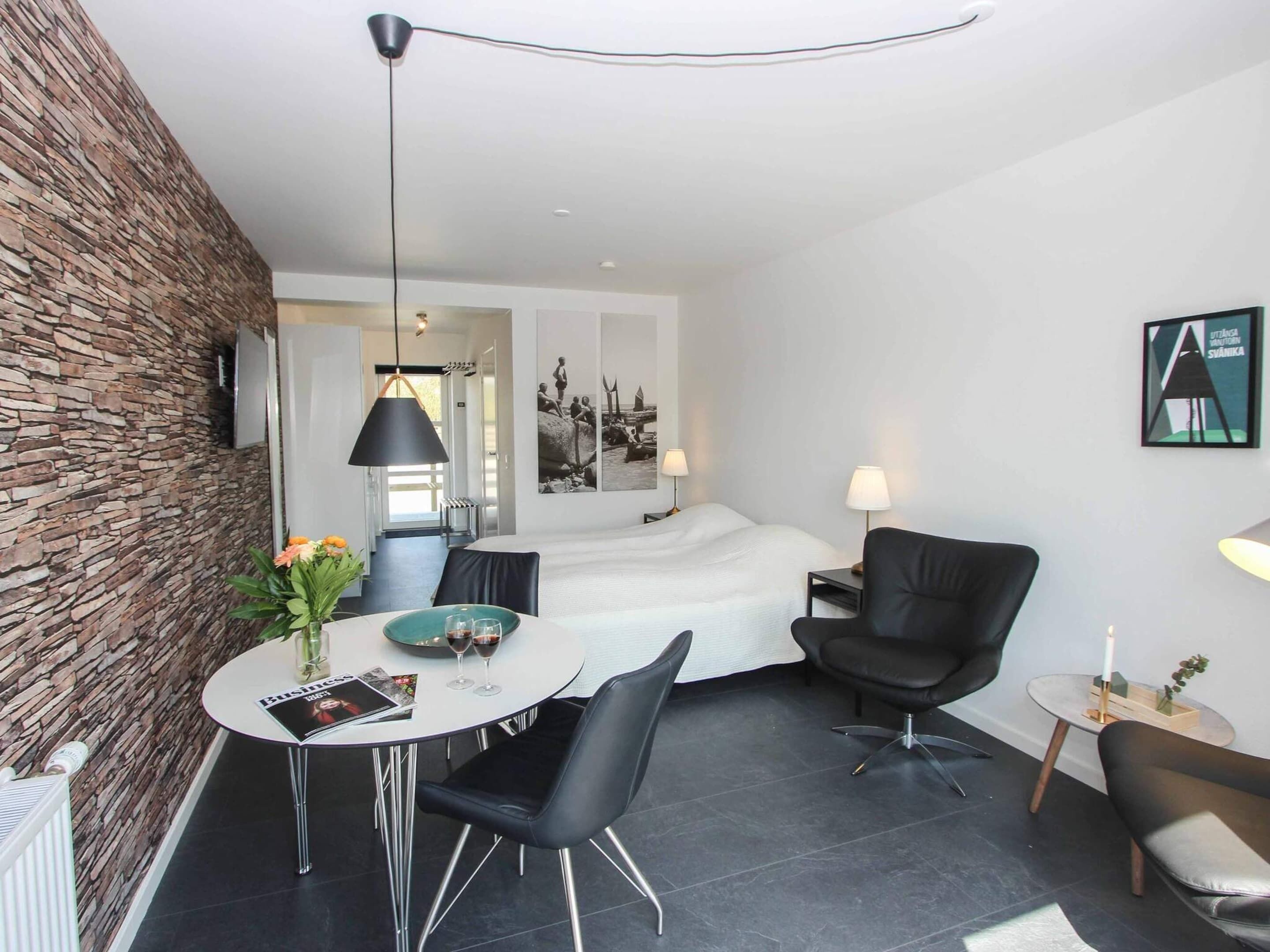 A modern hotel room with a stone accent wall, a bed, a dining table with chairs, a small side table, and a lamp.