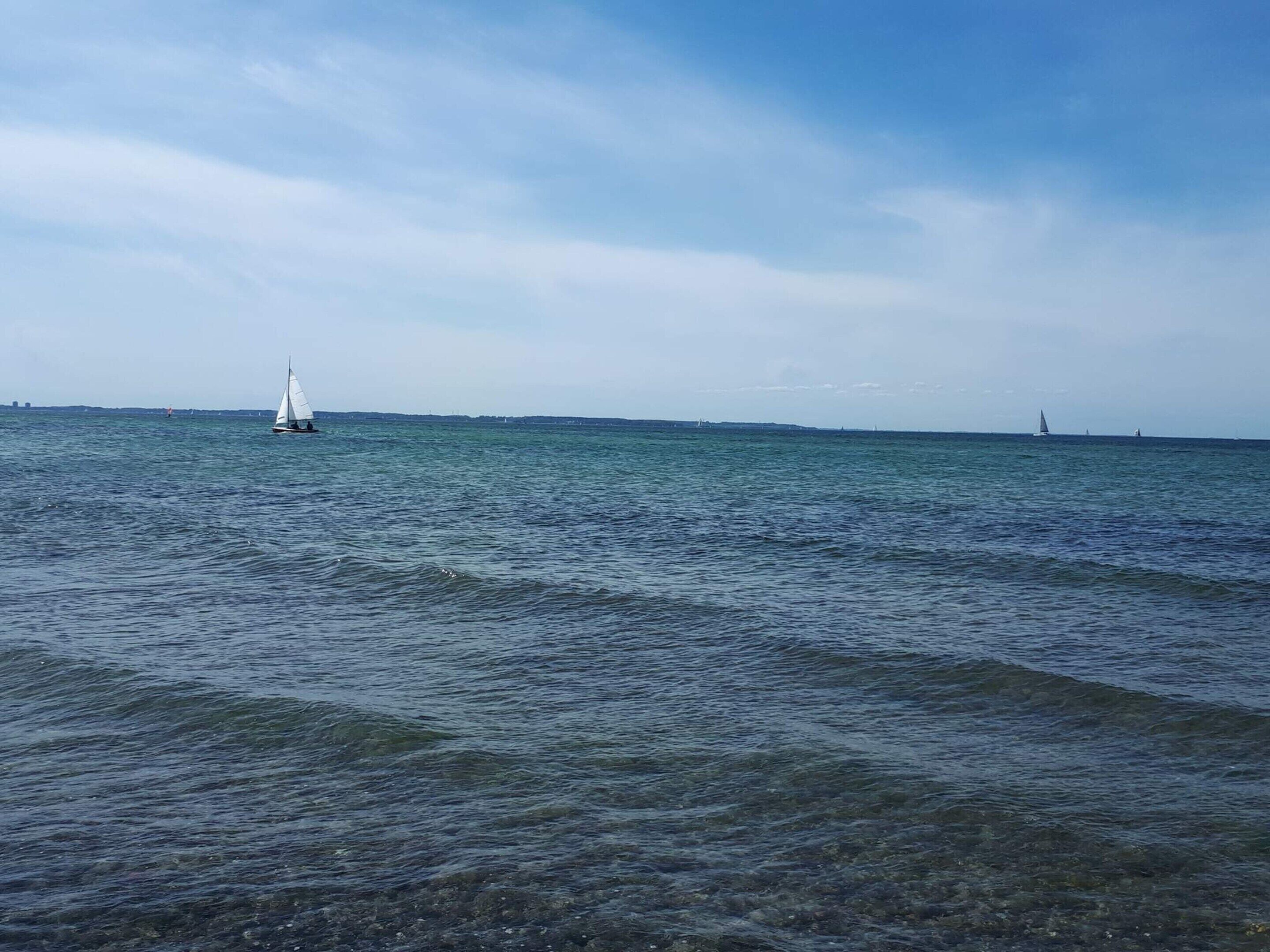 A calm sea with a sailboat in the distance and a lighthouse on the horizon.