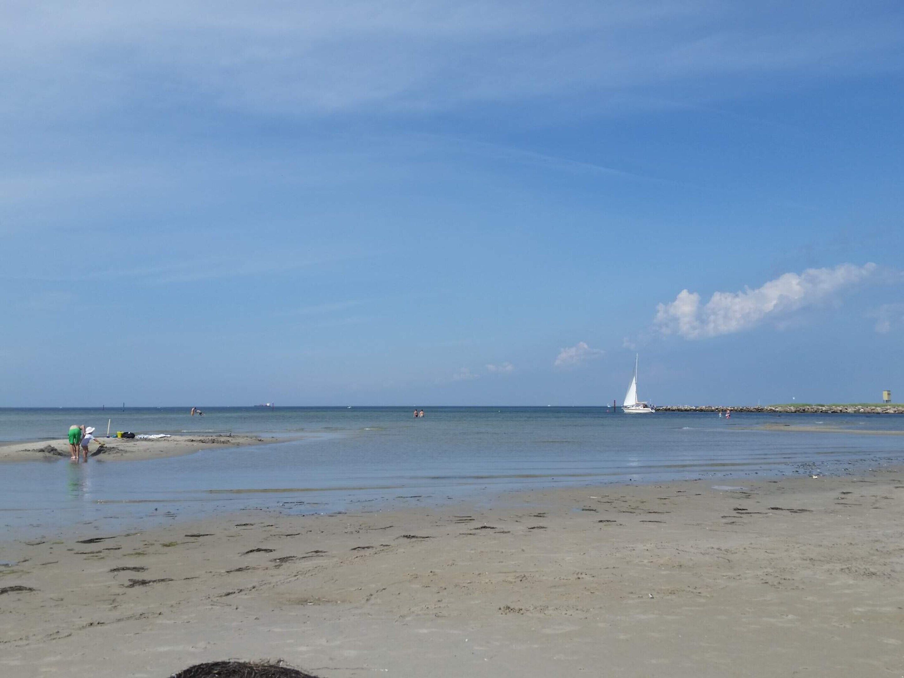 A beach with a sailboat in the distance, a rocky breakwater, and a few people near the water's edge.
