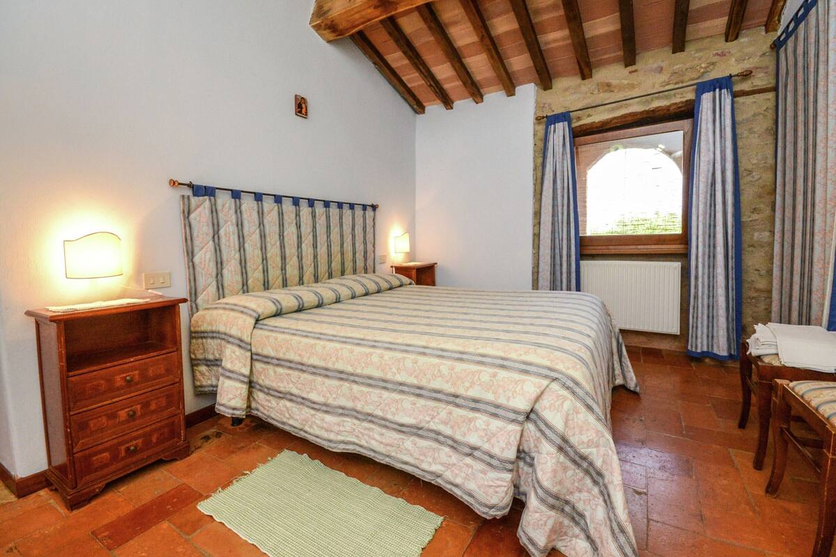 Ein Schlafzimmer mit Holzbalkendecke, einem Bett mit gestreifter Bettwäsche, einem Nachttisch mit Lampe, einer Kommode und einem Fenster mit Vorhängen.