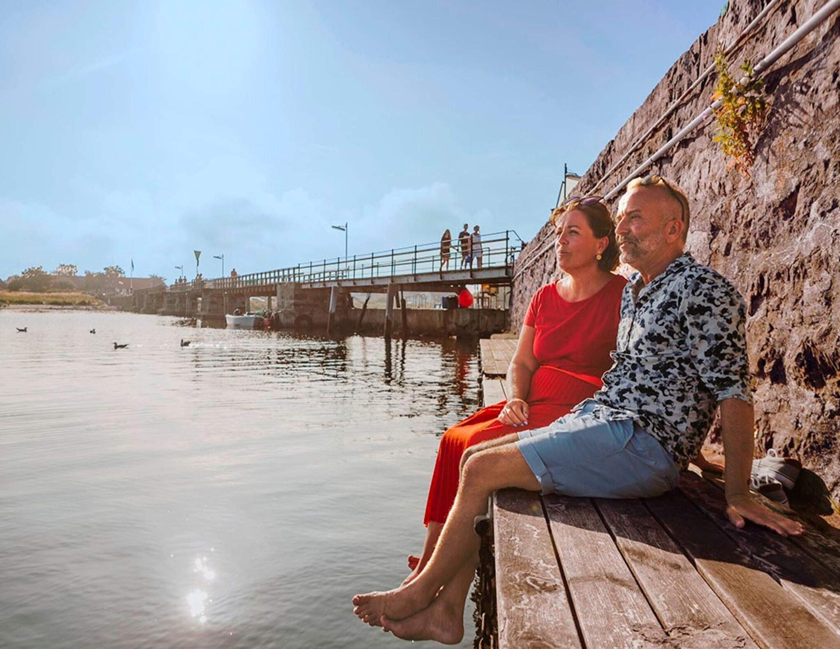 Un couple est assis sur un banc en bois, au bord de l’eau, et contemple un plan d’eau paisible où nagent des canards.