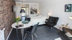 Une chambre d’hôtel moderne avec un mur en pierre, un lit, un coin repas avec une table et des chaises, et un petit coin salon avec un fauteuil et une lampe.