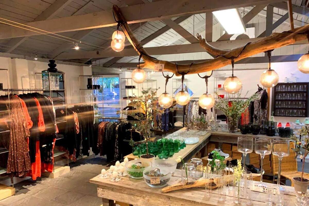 A retail space with clothing racks, a central display table with glassware, and hanging light fixtures.