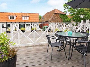 Une terrasse en bois avec une table ronde et quatre chaises, une clôture blanche et, au fond, des bâtiments aux toits recouverts de tuiles rouges.