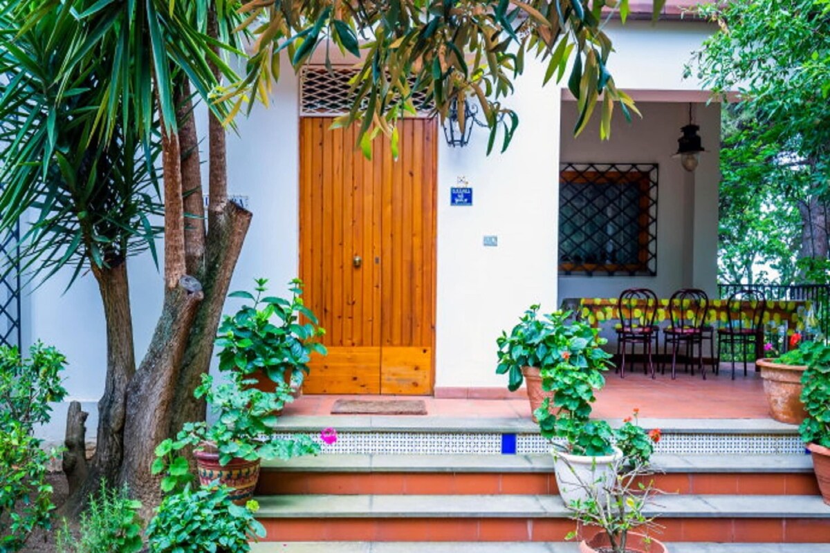 A wooden door with a sign above it, steps leading up to a porch with potted plants, and a table with chairs outside.