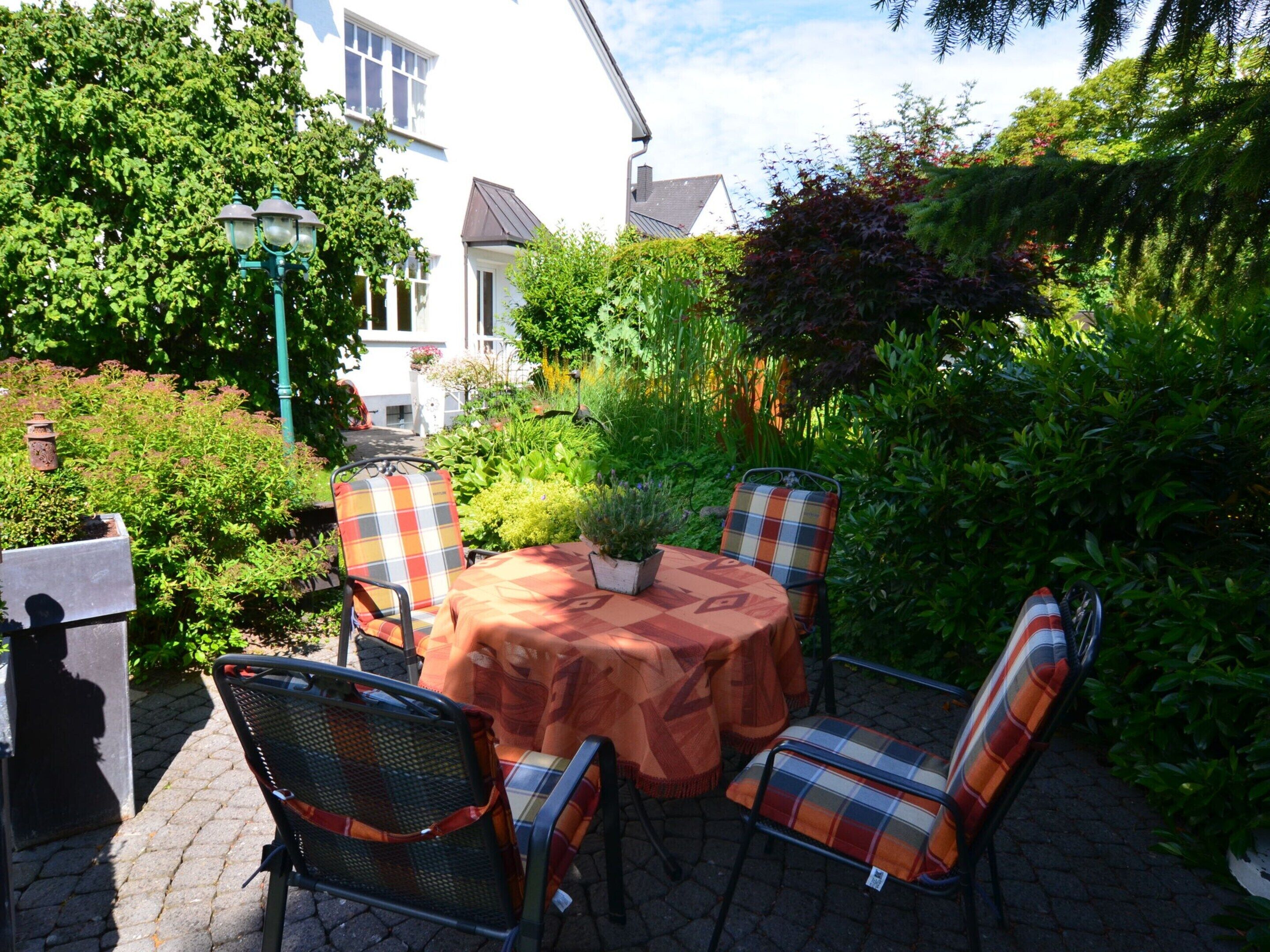 Une terrasse extérieure avec une table ronde et quatre chaises, entourée d’une végétation luxuriante et d’un bâtiment blanc en arrière-plan.