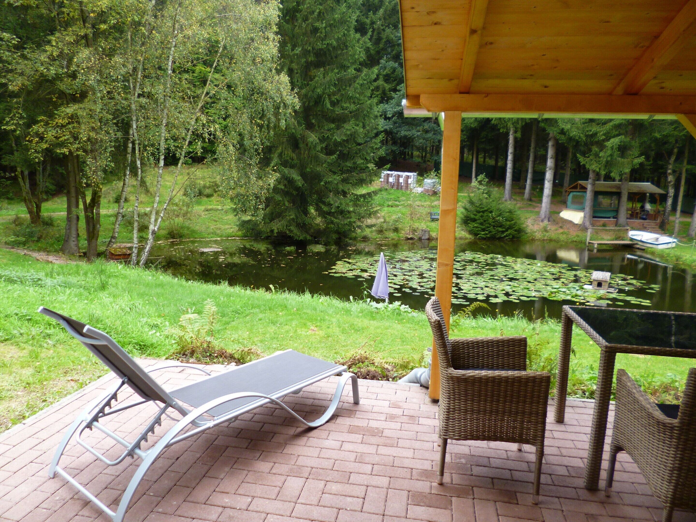 Eine Terrasse mit Liegestuhl und Korbstühlen mit Blick auf einen Teich mit Seerosen und einer Holzterrasse mit überdachtem Bereich.
