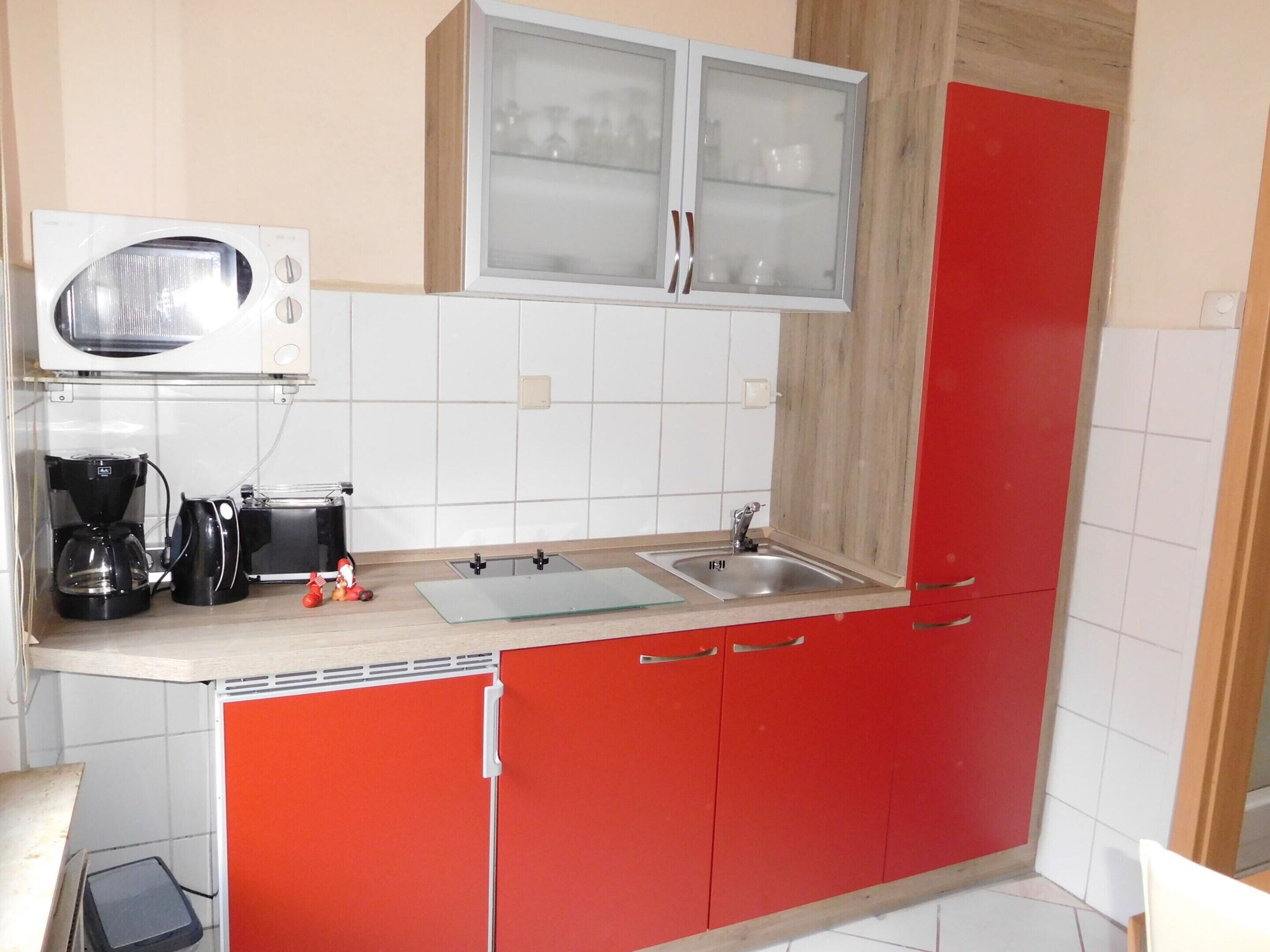 A compact kitchen with a microwave, coffee maker, and toaster. It features a sink, red cabinets, and a glass cabinet above the counter.