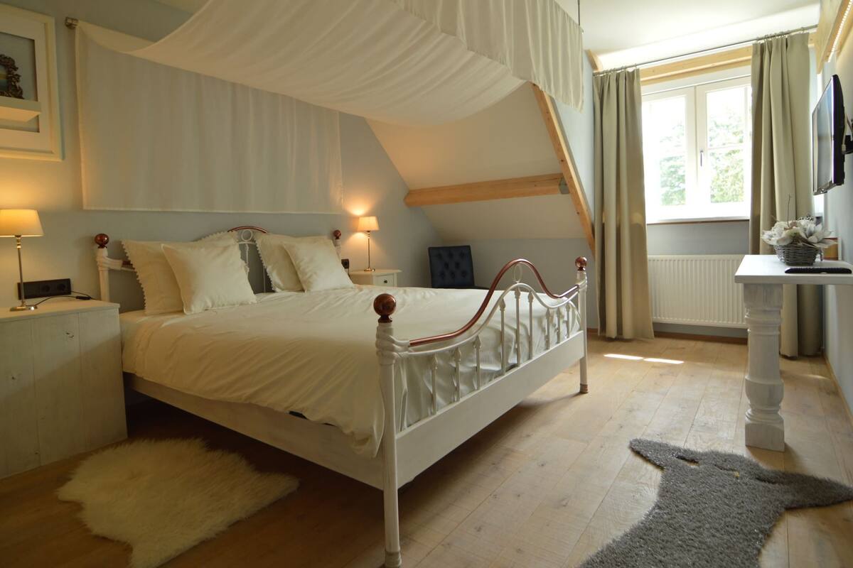 Une chambre à coucher comprenant un lit, une table de chevet, une lampe, une fenêtre avec des rideaux et une vue sur la verdure à l’extérieur.