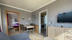 Una sala de estar moderna con un sofá, una mesa de comedor con sillas, un televisor montado en la pared y una cocina visible a través de un umbral abierto.