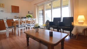 Un salon moderne avec une table basse en bois, des chaises blanches, un canapé noir et une table à manger avec des chaises. On y trouve une fenêtre avec des rideaux, une lampe et une petite plante sur la table basse.