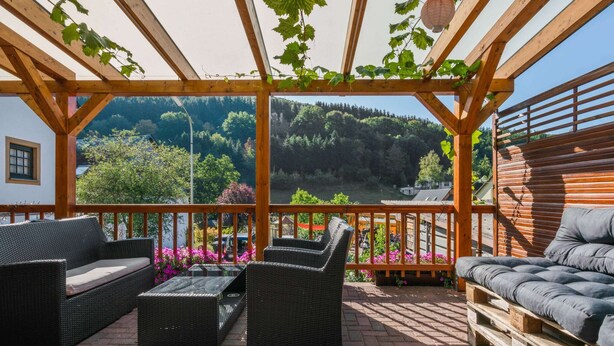 Et overdækket udendørs siddeområde med fletmøbler, et træpergola og udsigt til et bjerglandskab.