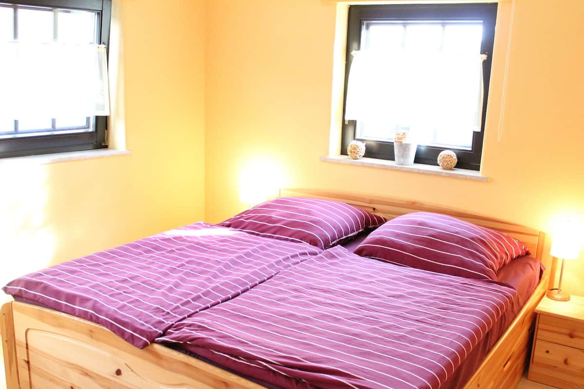 A bed with a wooden frame and purple bedding, a bedside table with a lamp, and a window with a view of the outside.
