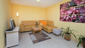 En stue med sofa, sofabord, en bærbar computer på et skrivebord, en potteplante og en væg med et blomsterbillede.