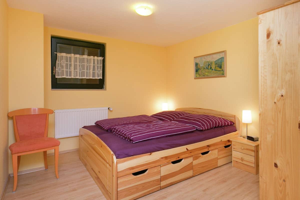 A bedroom with a wooden bed, a chair, a radiator, a window with a curtain, a painting on the wall, and a wooden wardrobe.