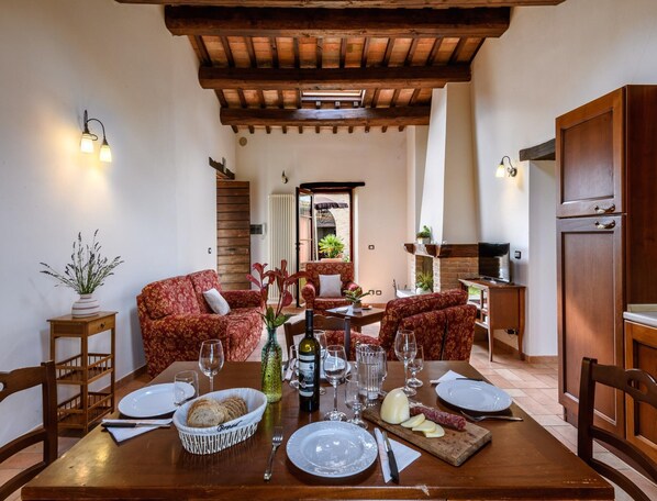 Un coin repas avec une table en bois dressée pour deux personnes, entouré de canapés rouges à motifs, d’une petite table d’appoint avec un vase, et donnant sur un espace cuisine.