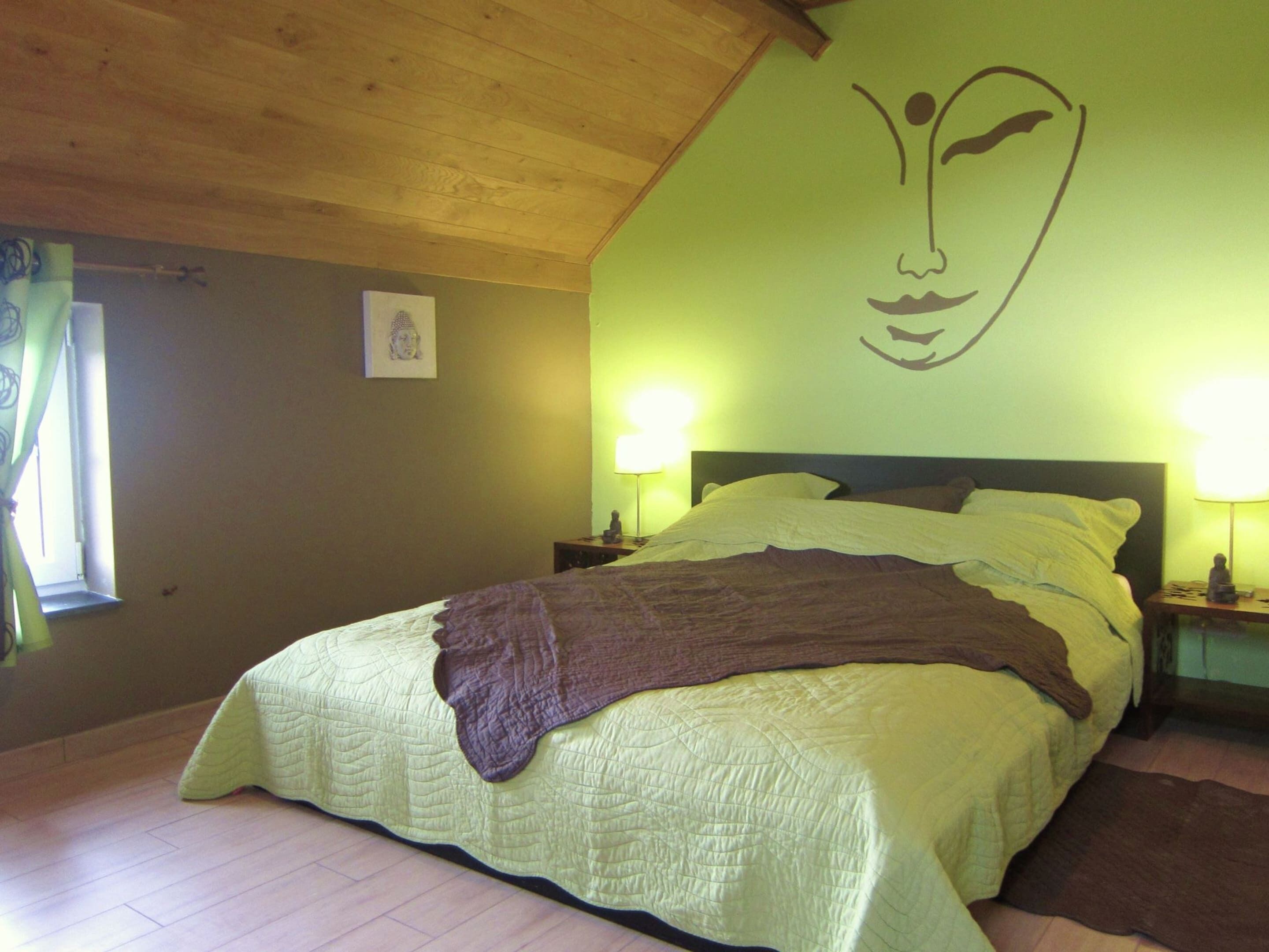 Ein Schlafzimmer mit einem Bett, zwei Nachttischlampen, einer an der Wand befestigten Buddha-Statue und einem Fenster mit Vorhängen.