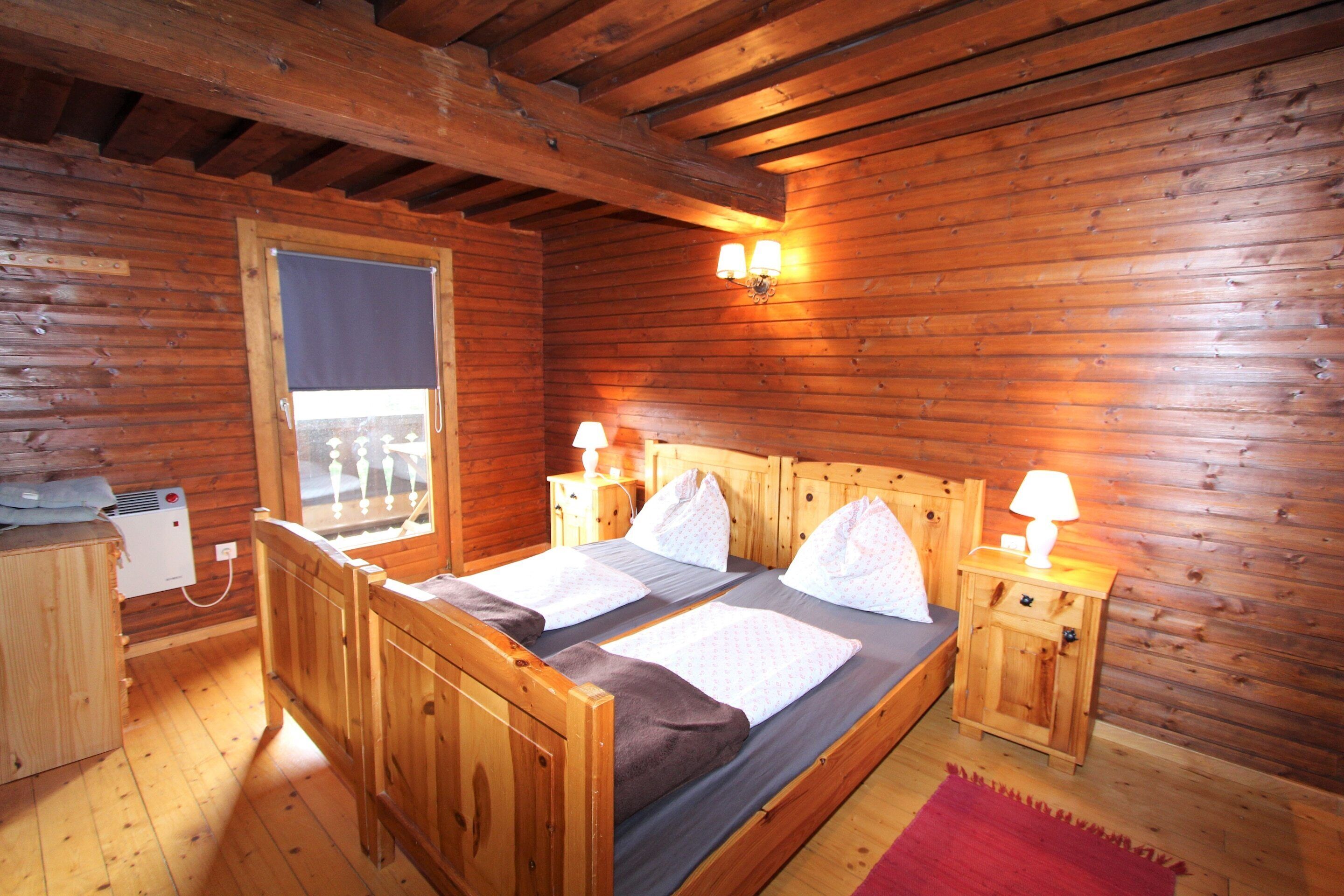 A wooden cabin room with a double bed, two bedside tables with lamps, a window with a view, and a heater.