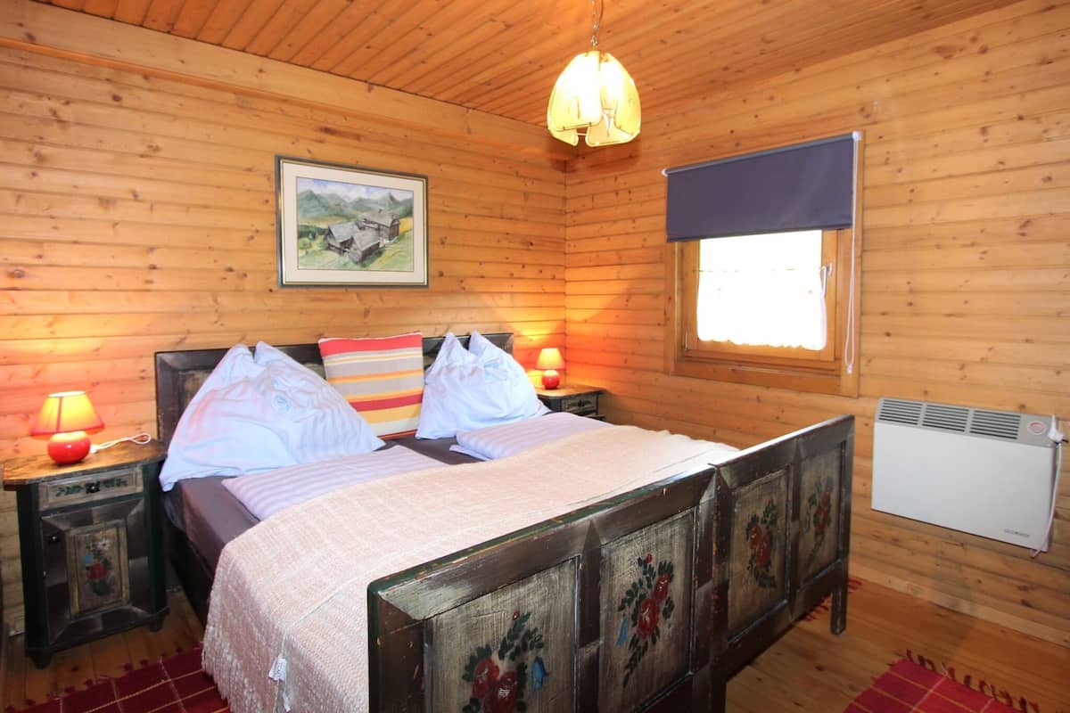 A wooden cabin bedroom with a bed, two bedside tables, a window with a blind, a hanging light fixture, and a radiator.