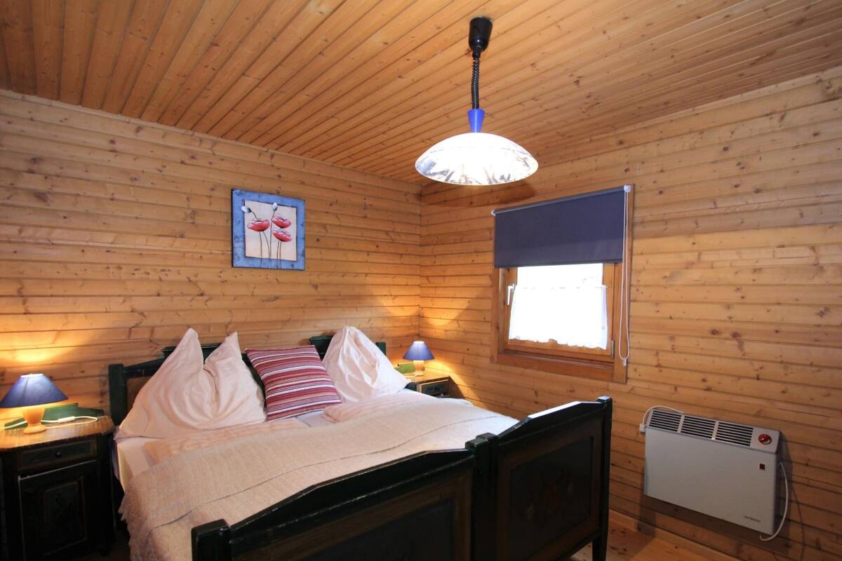 A wooden-paneled bedroom with a bed, bedside lamps, a window with a blind, and a wall-mounted heater.