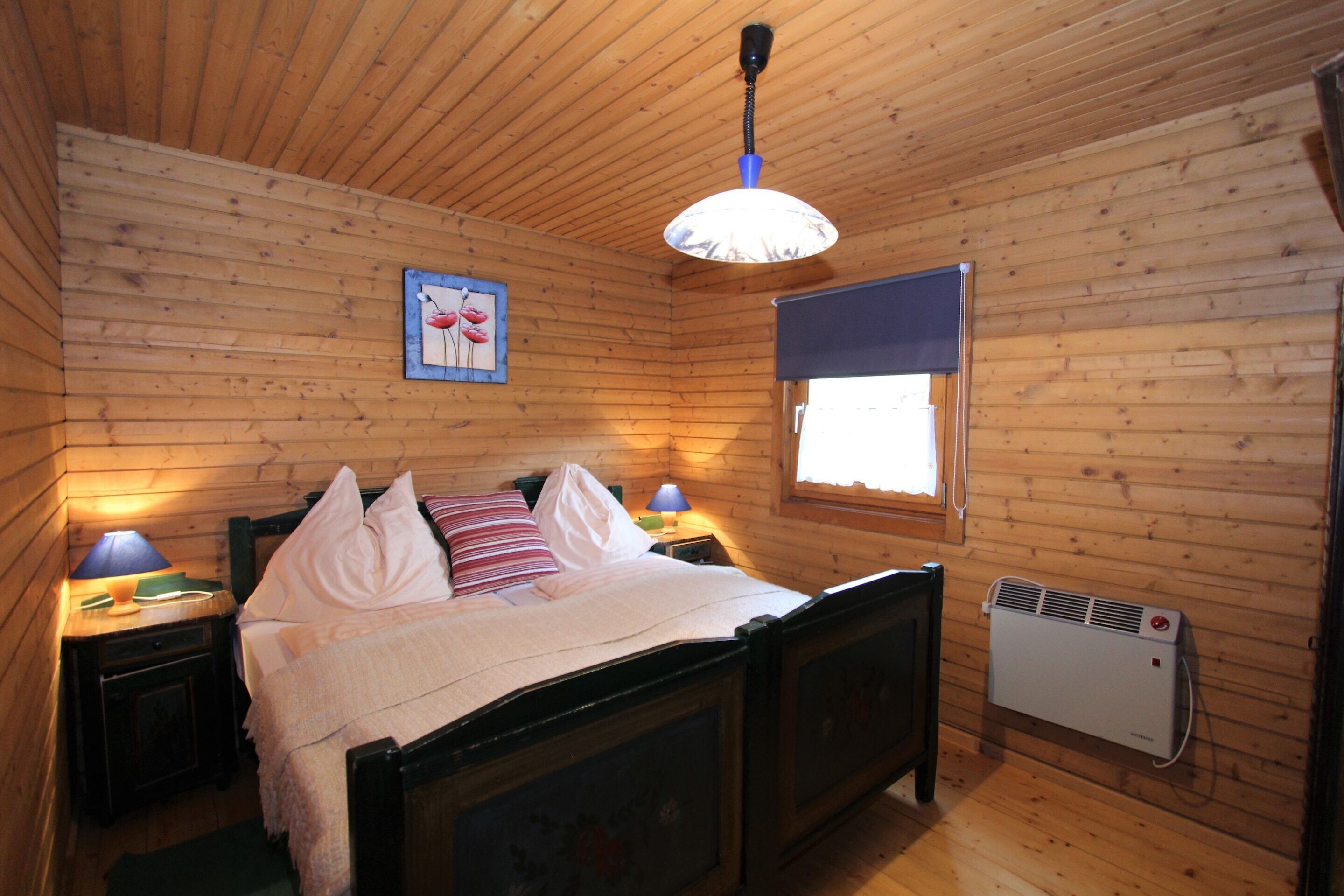 A wooden-paneled bedroom with a bed, bedside lamps, a window with a blind, and a wall-mounted heater.