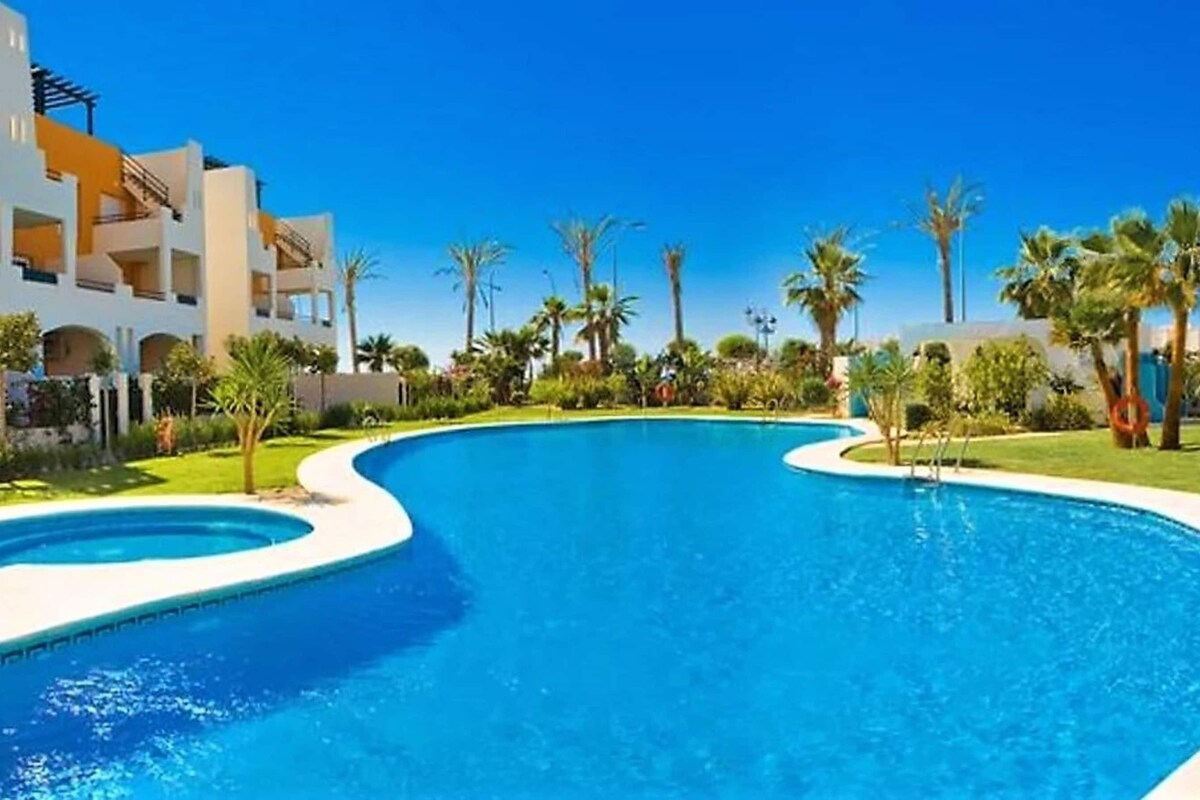 Una piscina con diseño curvo, rodeada de palmeras y un edificio de varios pisos con balcones.