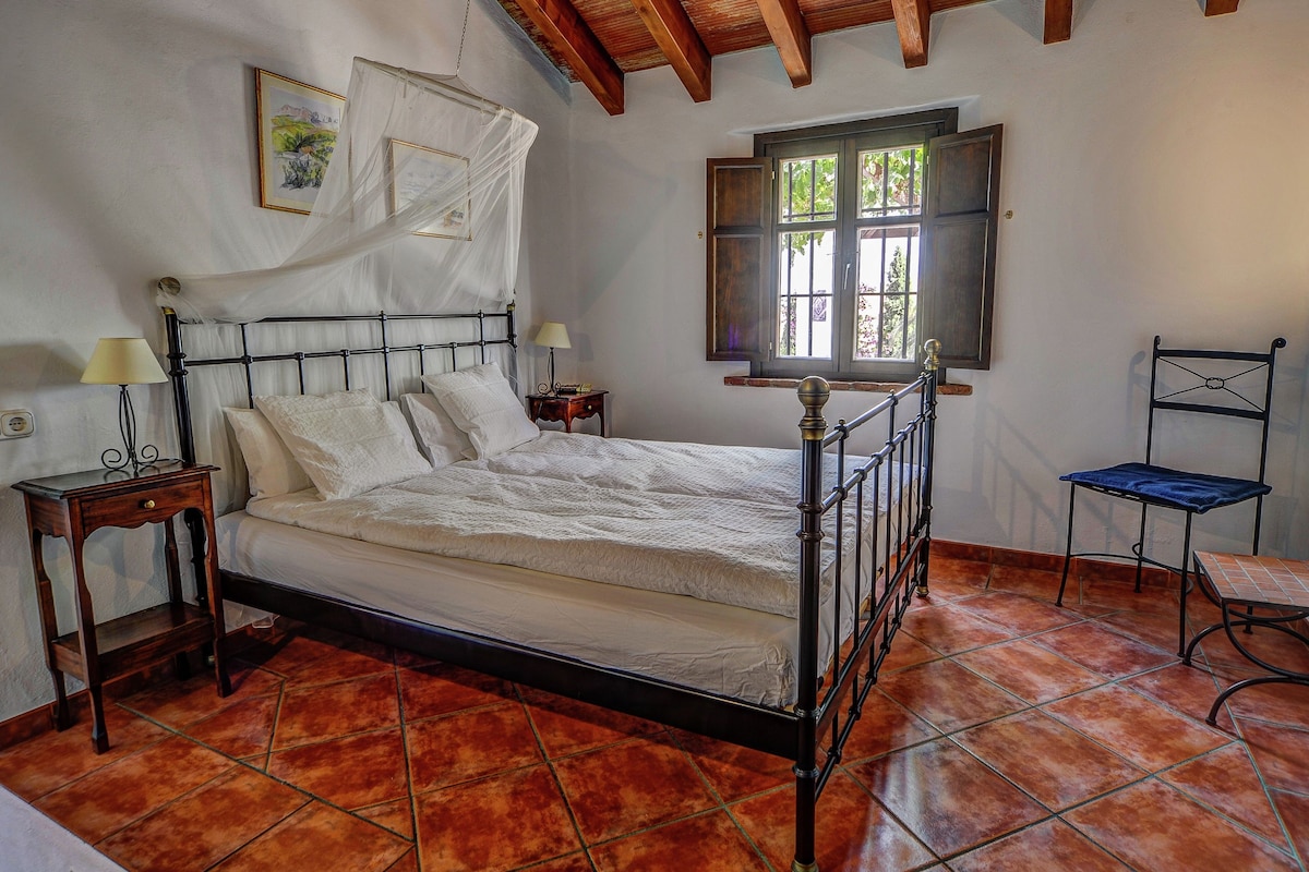 A bedroom with a bed, bedside tables, a chair, a window, and a wooden ceiling.