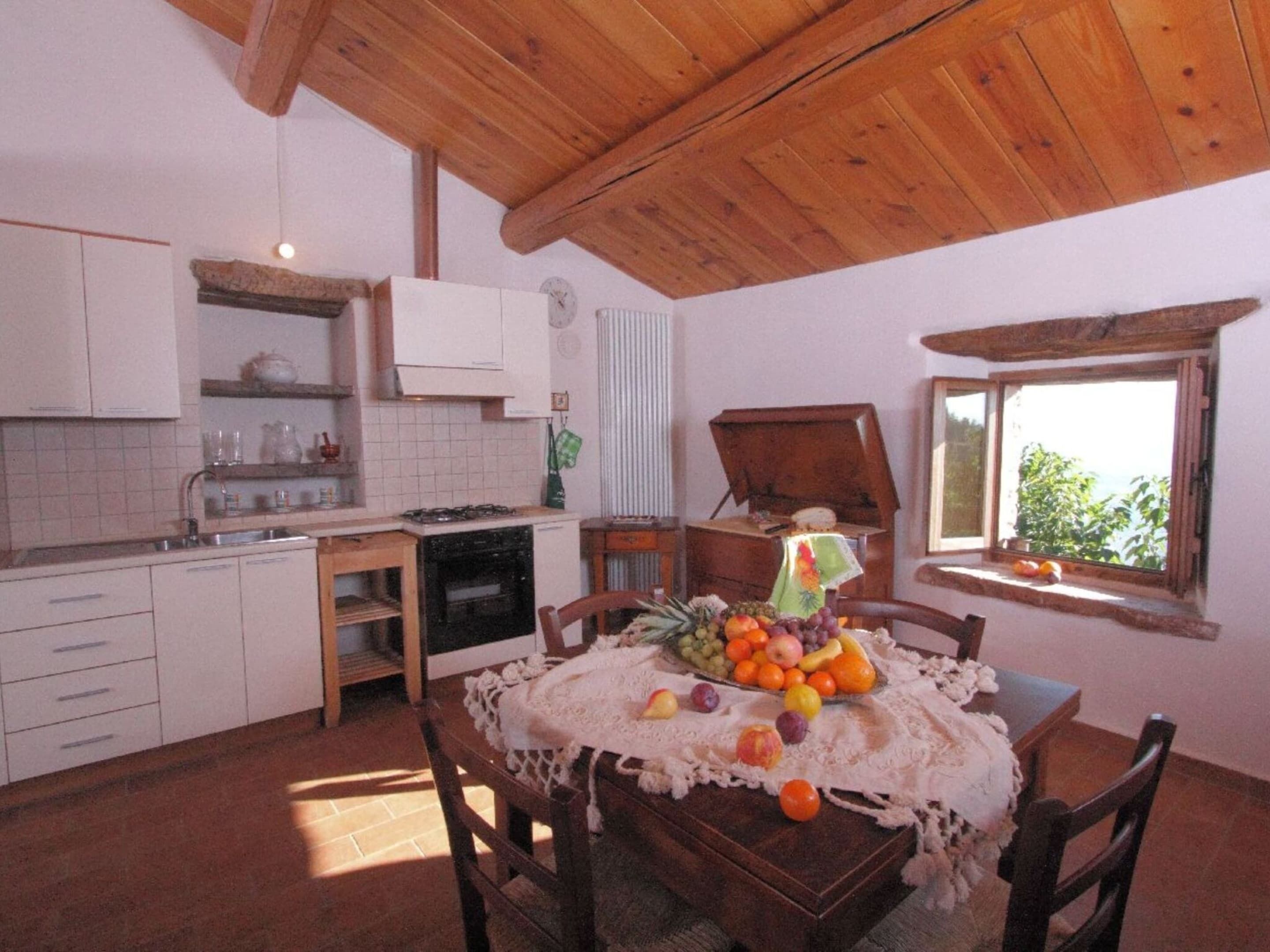 A rustic kitchen with white cabinets, a wooden dining table with a lace tablecloth, and a window with a view of greenery.
