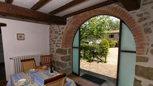 Un comedor con una mesa dispuesta para dos personas, una silla de mimbre y vistas a un patio exterior con una parrilla y follaje.